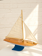 Load image into Gallery viewer, A wooden model sailboat with cream-colored sails, a polished wood hull, and a blue base, displayed against a white brick wall with warm sunlight casting shadows.