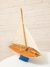 Load image into Gallery viewer, A wooden model sailboat with cream-colored sails, a light wood deck, and a blue hull, displayed on a white surface against a white brick wall background.