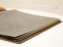 Load image into Gallery viewer, A close-up of a leather-bound book or journal with blank, textured gray pages, resting on a white textured fabric surface.