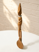 Load image into Gallery viewer, A tall, slender hand-carved wooden sculpture of a figure with a spoon-like form at the base, standing on a white textured surface against a white brick wall with soft window shadows.