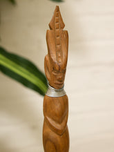 Load image into Gallery viewer, A close-up of a carved wooden decorative object with a stylized face and tall pointed top, featuring a silver metal band around the neck, set against a softly blurred indoor background with a green plant leaf visible on the left.