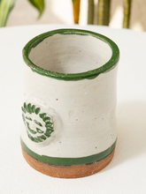 Load image into Gallery viewer, A handmade ceramic cup with a speckled off-white glaze, a dark green rim, a green decorative leaf motif, and a natural brown clay foot, shown on a white surface.
