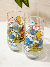 Load image into Gallery viewer, Two clear glass tumblers with colorful Smurf illustrations, displayed on a white textured surface against a light brick wall background.