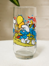Load image into Gallery viewer, A clear glass drinking tumbler featuring a colorful printed illustration of Smurf characters, shown on a white textured surface against a light beige background.