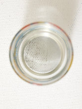 Load image into Gallery viewer, A top-down view of a clear glass jar or bottle with a colorful painted rim, showing a textured white interior surface with fine speckles, set against a white textured background.