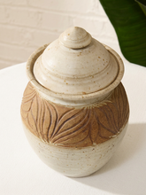 Load image into Gallery viewer, A handmade ceramic lidded jar with a speckled cream glaze and a brown textured band featuring embossed leaf patterns, set on a white surface against a white brick wall background.