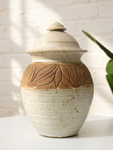 Load image into Gallery viewer, A handmade ceramic lidded jar with a textured speckled cream glaze and a contrasting brown band featuring leaf-like relief patterns, set against a white brick wall background.