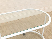 Load image into Gallery viewer, A close-up of a white metal coffee table with a curved glass top, set on a beige woven rug against a white brick wall.
