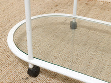 Load image into Gallery viewer, Close-up of a white metal rolling side table with a clear glass shelf and black caster wheels, shown on a beige woven rug.