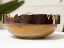 Load image into Gallery viewer, A handmade ceramic bowl with a glossy dark brown glazed rim featuring cream-colored drips, and a matte tan-brown glazed body, set against a soft white background with a hint of green foliage.