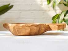 Load image into Gallery viewer, A carved wooden bowl with a rustic, hand-hammered texture and a small handle on one side, resting on a white textured surface with a white brick wall and green plants in the background.