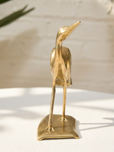 Load image into Gallery viewer, A small polished brass heron figurine standing on a rectangular base, photographed on a white surface against a softly blurred light background with a hint of greenery.