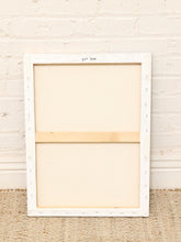Load image into Gallery viewer, An empty canvas print on a wooden frame, showing the back side with a wooden support bar, leaning against a white brick wall on a woven jute rug.