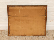 Load image into Gallery viewer, The back of a vintage wooden picture frame, showing the cardboard backing, mounting pins, and hanging wire, set against a white brick wall and woven rug.