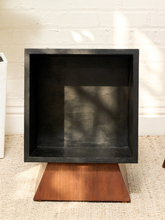 Load image into Gallery viewer, A square black metal display shelf or mount with a triangular wooden base, set against a white brick wall on a textured beige rug.