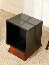 Load image into Gallery viewer, A modern cube-shaped side table with a dark gray glossy top and a warm brown wood base, set on a woven jute rug.
