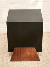 Load image into Gallery viewer, A matte black cube side table or stool with a triangular wooden footrest, set on a beige woven rug against a white brick wall.