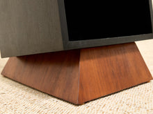 Load image into Gallery viewer, Close-up of a modern TV stand or console base made of warm medium-brown wood grain, supporting a dark gray or black television on a textured beige carpet.