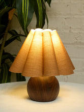 Load image into Gallery viewer, A vintage-style table lamp with a pleated beige fabric shade and a dark brown wooden base, illuminated on a white surface with a white brick wall and green plant leaves in the background.