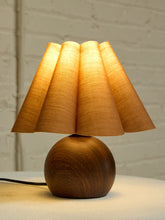 Load image into Gallery viewer, A table lamp with a warm glowing pleated fabric shade and a round dark wood base, set on a light surface against a white brick wall background.