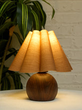 Load image into Gallery viewer, A table lamp with a pleated beige fabric shade and a round dark wood base, lit warmly against a white brick wall with a green plant in the background.