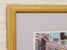 Load image into Gallery viewer, A close-up of a framed art print showing a yellow-gold frame, a light lavender mat, and a watercolor painting of brick houses with red roofs against a white brick wall background.