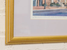 Load image into Gallery viewer, A close-up of a framed art print showing the corner of a gold frame, white mat board, and a watercolor painting of a street scene with a signature at the bottom.