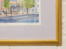 Load image into Gallery viewer, A framed watercolor painting of a street scene with trees and buildings, mounted with a wide light lavender mat and enclosed in a gold frame against a white textured wall.