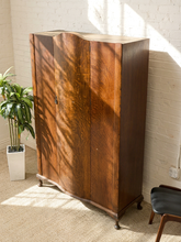 Load image into Gallery viewer, A tall wooden cabinet with two doors and curved top, photographed in a room corner against a white brick wall with sunlight and leaf shadows. A potted plant is on the left and part of a chair is visible on the right.