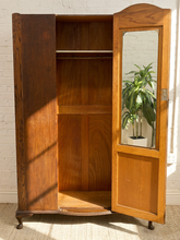 Load image into Gallery viewer, A vintage wooden armoire cabinet with one door open, showing its interior shelves and wood grain. The door features an arched top and a rectangular mirror inset, reflecting a green potted plant.