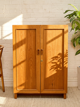 Load image into Gallery viewer, A two-door wooden cabinet with a warm honey finish and small brass handles, set against a white brick wall with sunlight casting leaf shadows on its doors.