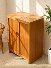 Load image into Gallery viewer, A mid-century modern wooden cabinet with two doors and a hinged top section, shown in a warm natural wood finish in a room corner with a white brick wall, beige carpet, and a plant.