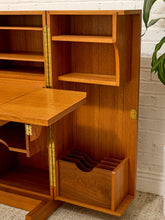 Load image into Gallery viewer, A mid-century modern wooden writing desk and cabinet unit with a warm honey finish, featuring brass hinges, a pull-out desktop, open shelving, and a lower organizer drawer, set against a white brick wall.