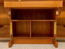 Load image into Gallery viewer, Close-up of a wooden cabinet or sideboard with two doors open, showing brass hinges and an open lower drawer. The wood has a warm medium brown finish, and the cabinet is set on a light beige textured carpet.