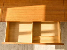 Load image into Gallery viewer, An open wooden drawer with a warm honey finish, showing a light wood divider and pull-out organizer tray inside, set against a textured beige rug with natural light casting shadows.