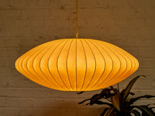 Load image into Gallery viewer, A warm, glowing oval pendant light with a pleated paper or fabric shade, hanging against a textured brick wall with a plant visible in the lower right corner.