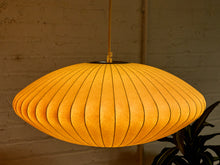 Load image into Gallery viewer, A mid-century modern pleated paper pendant light glowing with a warm golden-yellow light, hanging against a rustic brick wall background.