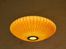 Load image into Gallery viewer, A warm, glowing paper lantern pendant light hanging from a thin cord against a textured brick wall.