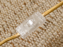 Load image into Gallery viewer, A close-up of a clear plastic inline switch or connector mounted on a gold braided cord, set against a textured beige woven background.
