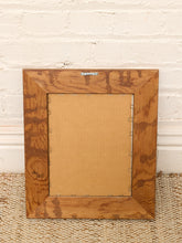 Load image into Gallery viewer, A square wooden picture frame with a visible grain pattern, shown from the back with its cardboard mounting insert, placed on a woven rug against a white brick wall.