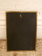 Load image into Gallery viewer, The back of a rectangular black picture frame with a gold-toned metal border and a hanging loop at the top, resting on a woven rug against a textured off-white wall.