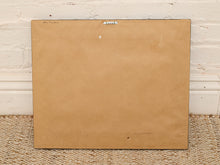 Load image into Gallery viewer, The back of a square framed artwork or print, showing a plain tan backing board with a metal hanger at the top, leaning against a white painted brick wall on a woven rug.