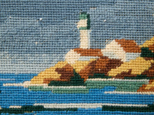 Load image into Gallery viewer, Close-up of a woven or needlepoint-style coastal scene with blue water, a white lighthouse, cream buildings, and tan/gold rocks under a blue sky.