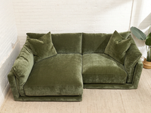 Load image into Gallery viewer, An olive green velvet sectional sofa with matching throw pillows, positioned in a room with a white brick wall and a light-colored rug.