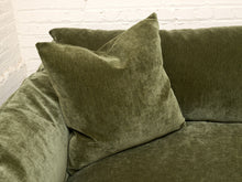 Load image into Gallery viewer, An olive green velvet throw pillow resting on an olive green velvet sofa in front of a white brick wall.