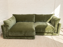 Load image into Gallery viewer, An olive green velvet sectional sofa with a chaise lounge on the left, positioned against a white brick wall on a light beige textured rug.