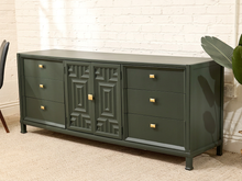 Load image into Gallery viewer, A dark green sideboard cabinet with gold hardware, featuring three drawers on each side and a central double-door panel with geometric detailing, set against a white brick wall and beige carpet.