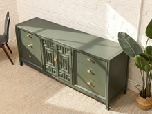 Load image into Gallery viewer, An olive green sideboard cabinet with gold hardware, styled in a room setting with a white brick wall, beige carpet, and a potted plant.