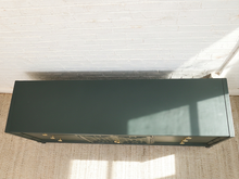 Load image into Gallery viewer, A dark green wooden cabinet with gold drawer pulls, photographed from a high angle against a white brick wall and beige rug.