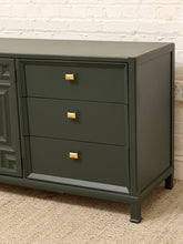 Load image into Gallery viewer, A dark green three-drawer dresser with small gold square knobs, shown in a room setting against a white brick wall and beige carpet.